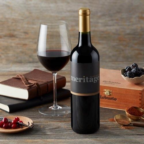 Okanagan Meritage Wine in a glass on a table beside a wine bottle sporting the Okanagan Meritage label. The wine is on a table beside berries, leather-bound books, and a small wooden case